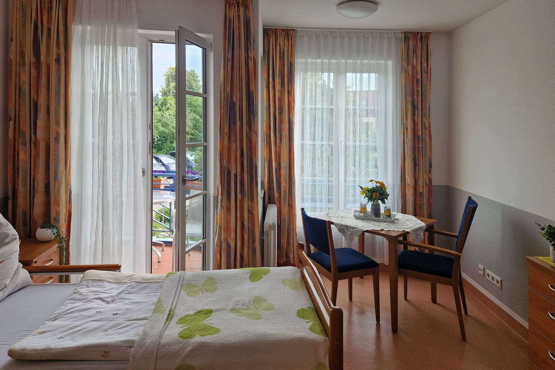 Zimmer mit Bett, Tisch und zwei Stühlen, die Balkontür ist offen. Blick zum Balkon und Fenster.
