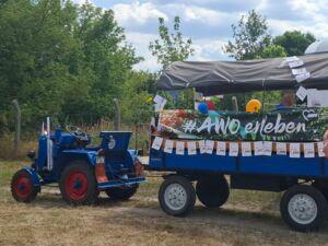 Von einem blauen Traktor gezogener blauer Kremser-Anhänger mit grauer Plane, geschmückt mit Luftballons und weißen AWO Wimpeln. Traktor ohne Fahrer.