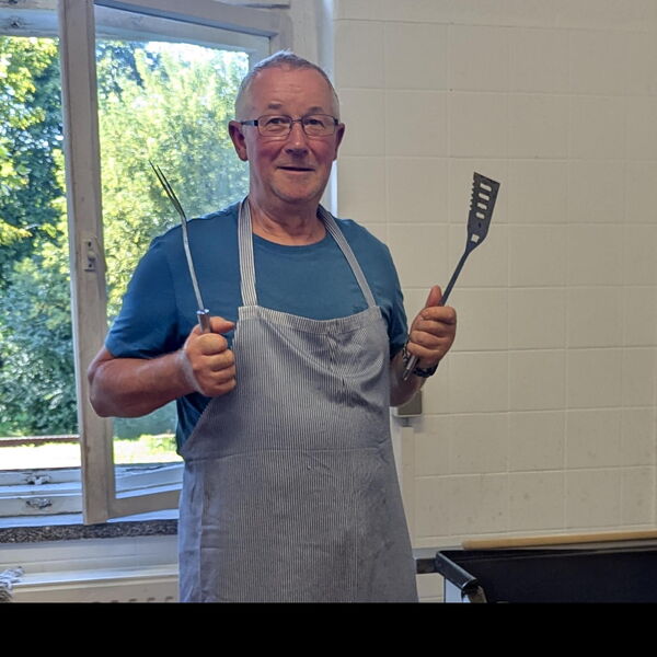 Mann mit Brille, Grillschürze und Grillbesteck in jeder Hand.