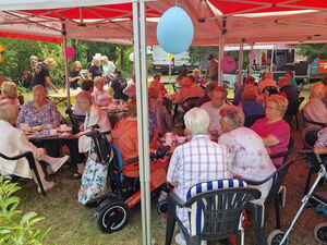 Fest. Viele ältere Menschen sitzen an Kaffeetischen unter mit Luftballons geschmückten Pavillons.