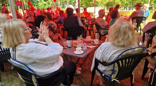 Menschen sitzen an kleinen Gartentischen bei Kaffee und Kuchen und klatschen.