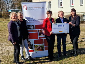 Draußen, Sonne scheint. Mittig ein Roll-Up von „Hilfe für Familien in Not“. Links davon zwei lächelnde Frauen. Rechts davon drei Frauen halten Scheck in die Kamera.