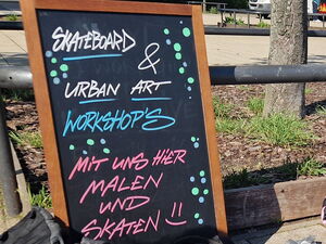 Draußen. Ein Schild: Skatebord und Urban Art Workshops.  3 Skateboards lehnen an einer Stand und Schutzkleidung wie Knieschützer liegen wild verteilt auf dem Boden,