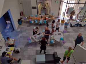 Im Museum. Großer Raum, von oben, durch die Eingangstür fällt Sonnenlicht. Viele Tische mit blauen und lila Luftballons geschmückt. Es gibt ein Büfett.