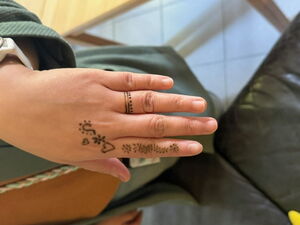 Hand mit Henna-Muster, bestehend aus zwei Herzen und kleinen Ornamenten auf Fingern und Handrücken, im Hintergrund Stoff und Fliesenboden.