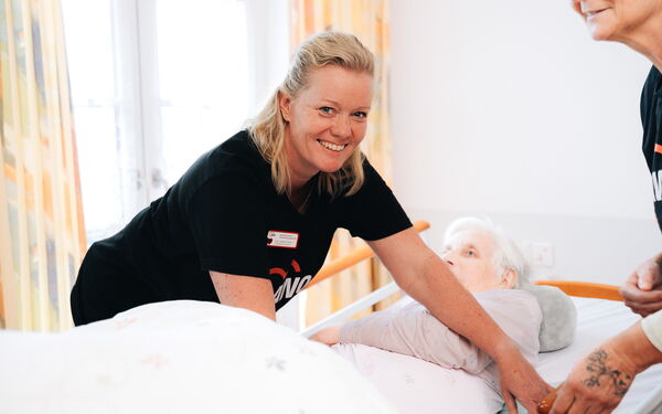 Eine Frau mit einem schwarzen AWO T-Shirt kümmert sich um eine alte Frau in einem Pflegebett und lächelt in die Kamera.