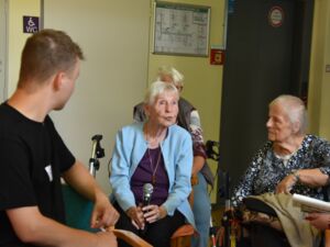 Alte Frau mit weißen Haaren sitzt auf einem Stuhl und hält ein Mikrofon mit beiden Händen die sie im Schoß abgelegt hat. Frau trägt hellblaue Strickjacke. Zwei junger Männer schauen interessiert zu ihr. Zwei weitere alte Frauen sitzen in der Nähne.