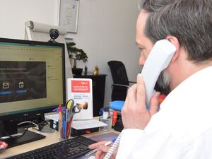 Mann an einem Schreibtisch am PC mit Telefonhörer am Ohr und Stift in der anderen Hand. Mann hat graue Haare und Bart, er trägt ein weißes langärmliches Hemd.