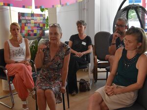4 Frauen, ein Mann sitzen auf Stühlen die in Kreisen angeornet sind. Blonde Frau mit einer Brille auf den Kopf geschoben spricht. Sie trägt ein geblümtes Kleid. Die anderen hören ihr zu.