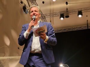 Mann in blauen Anzug und weißem Hemd steht in einem Raum. Er hat Mikro und Moderationskarten in der Hand. Im Hintergrund Stühle und Lampen.