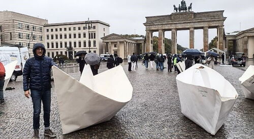 Vorplatz des Brandenburger Tor in Berlin. Regenwetter. Menschen mit Regenschirmen und Kapuzenjacken. 2 XXL-Origami-Papierboote stehen auf dem Platz. Ein Mann steht neben einem der Boote, er trägt eine dunkelblaue Regenjacke die Kapuze hat er aufgesetzt.