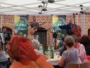 Eine Band aus drei Männer mit Akkordeon, Gitarre und Chello auf einer kleinen Bühne. Davor sitzen Menschen, größtenteils Frauen.