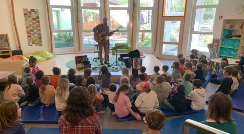 Mann mit Gitarre und Mikrofonständer steht vor einen großen Fenster. Publikum Kinder sitzen davor.