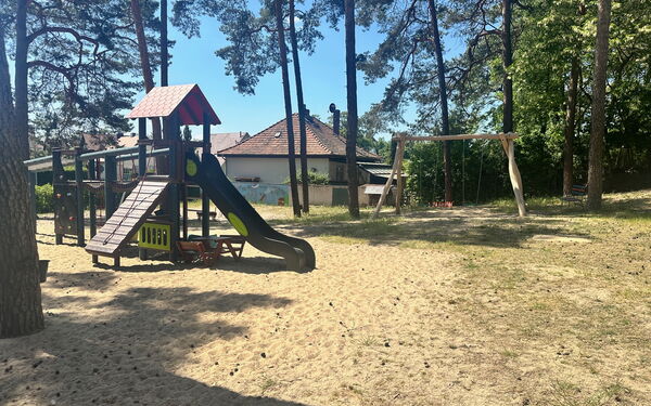 Draußen, Sonne scheint, Bäume sorgen für Schatten. Spielplatz. Schaukeln und ein Klettergerüst mit Rutsche.