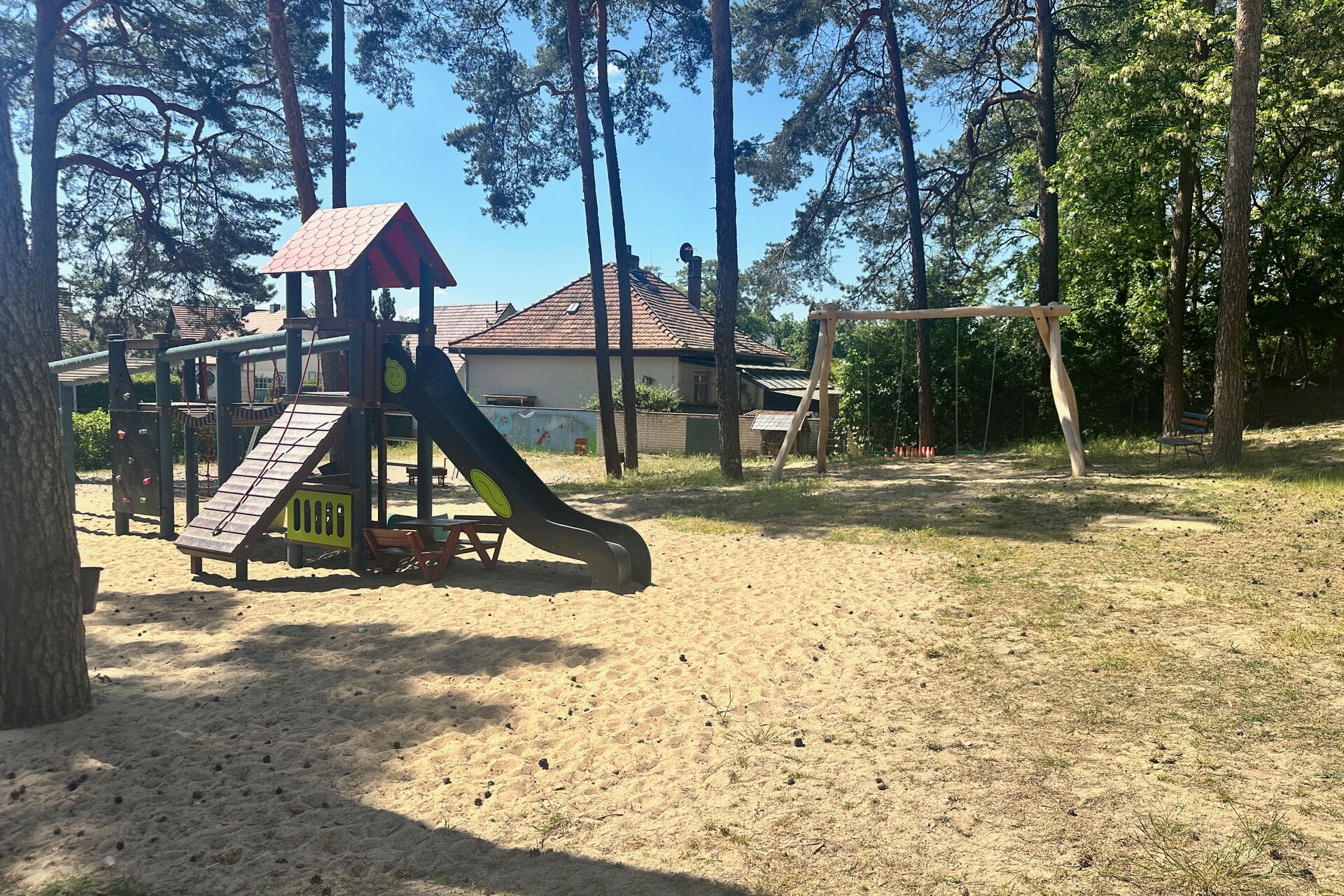 Draußen, Sonne scheint, Bäume sorgen für Schatten. Spielplatz. Schaukeln und ein Klettergerüst mit Rutsche.