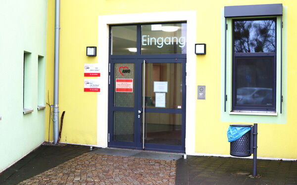 Eingangstür ist geschlossen. AWO Logo an der Glastür. Das Gebäude ist gelb. Im Fenster spiegelt sich ein Auto. Ein Mülleiner ist Eingangsbereich.