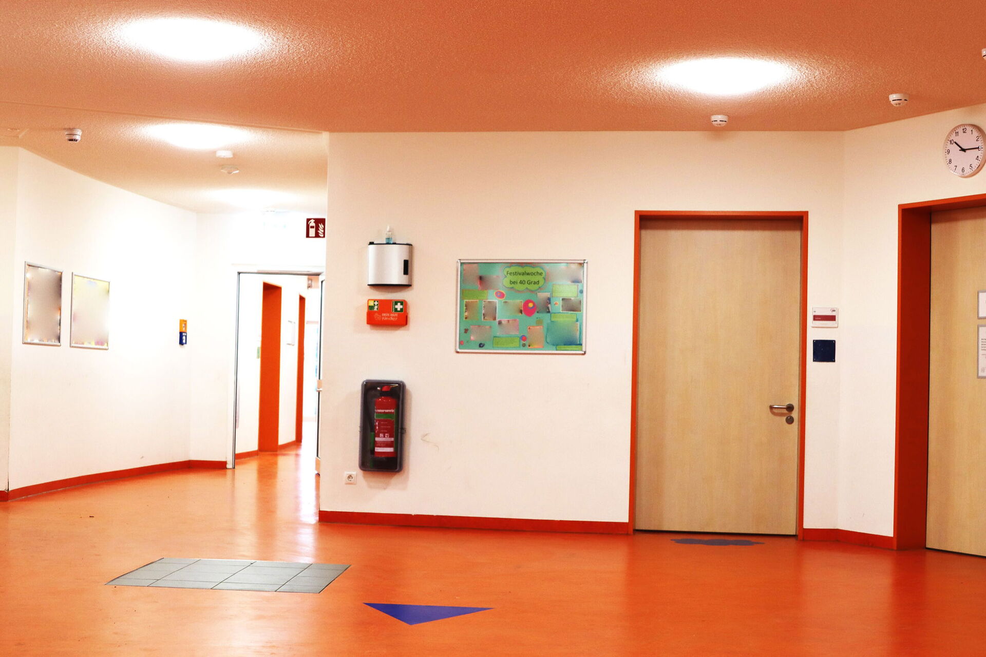 Der Flur ist breit. Der Boden ist orange, es befinden sich Markierungen auf dem Boden, die Wände haben eine helle Farbe.