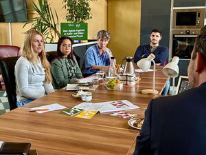 Heller Raum, Wohn-Küche. 5 Menschen an einem Tisch. Ein Mann nur von hinten. Die Anderen im Gespräch mit ihm, ihm gegenüber.