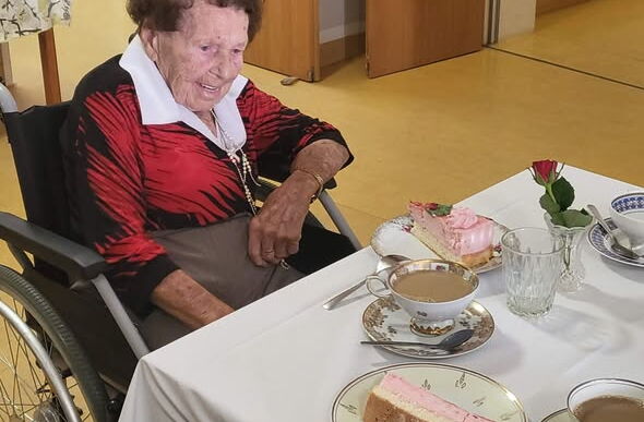 Seniorin im Rollstuhl am gedeckten Kaffeetisch. Nostalgisches Geschirr, ein großes Tortenstück und Kaffee stehen vor ihr.
