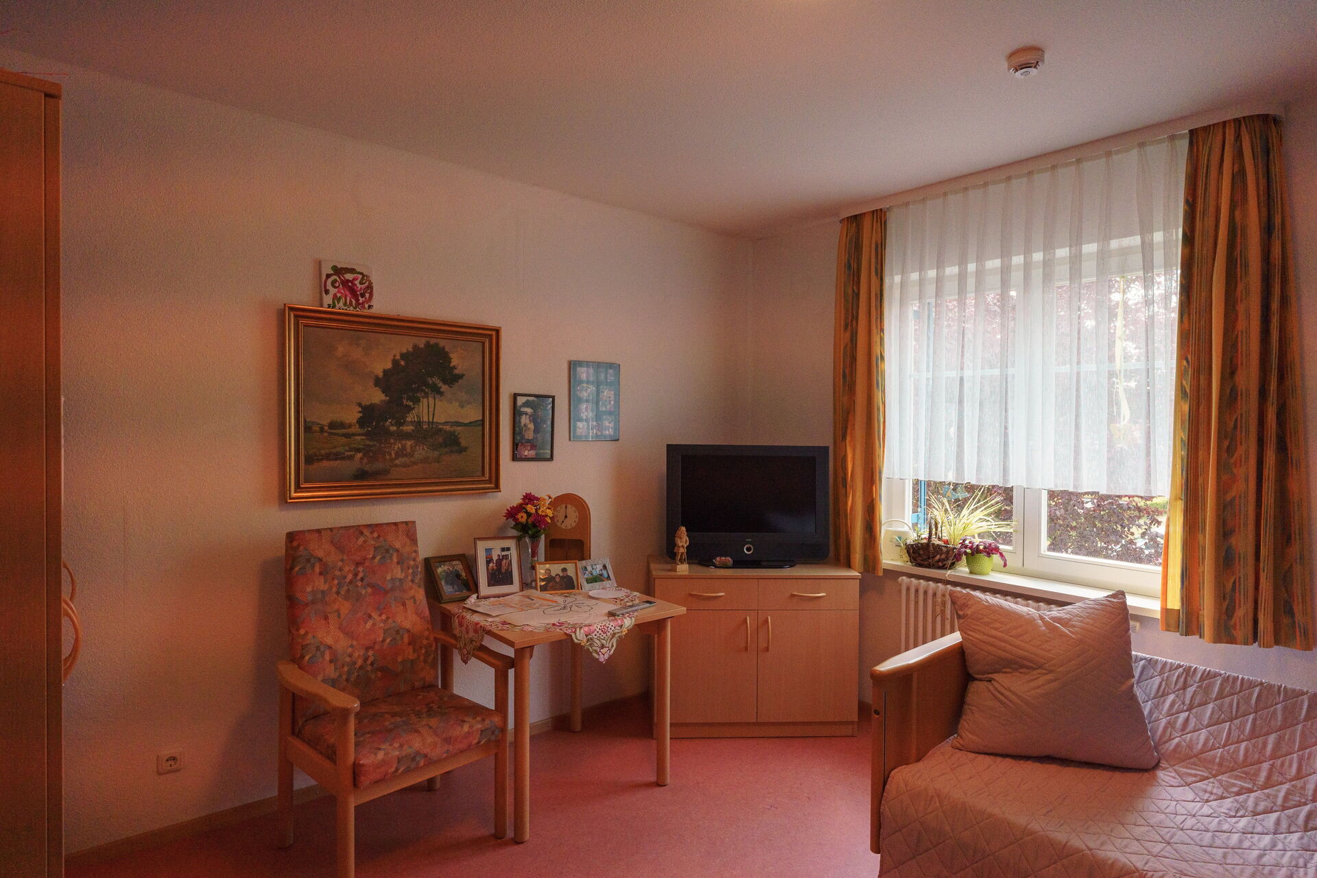Zimmer. Großes Fenster rechts, Fernseher auf Kommode in der Ecke, kleiner Tisch und Stuhl daneben. Über dem Tisch ein Landschaftsbild.