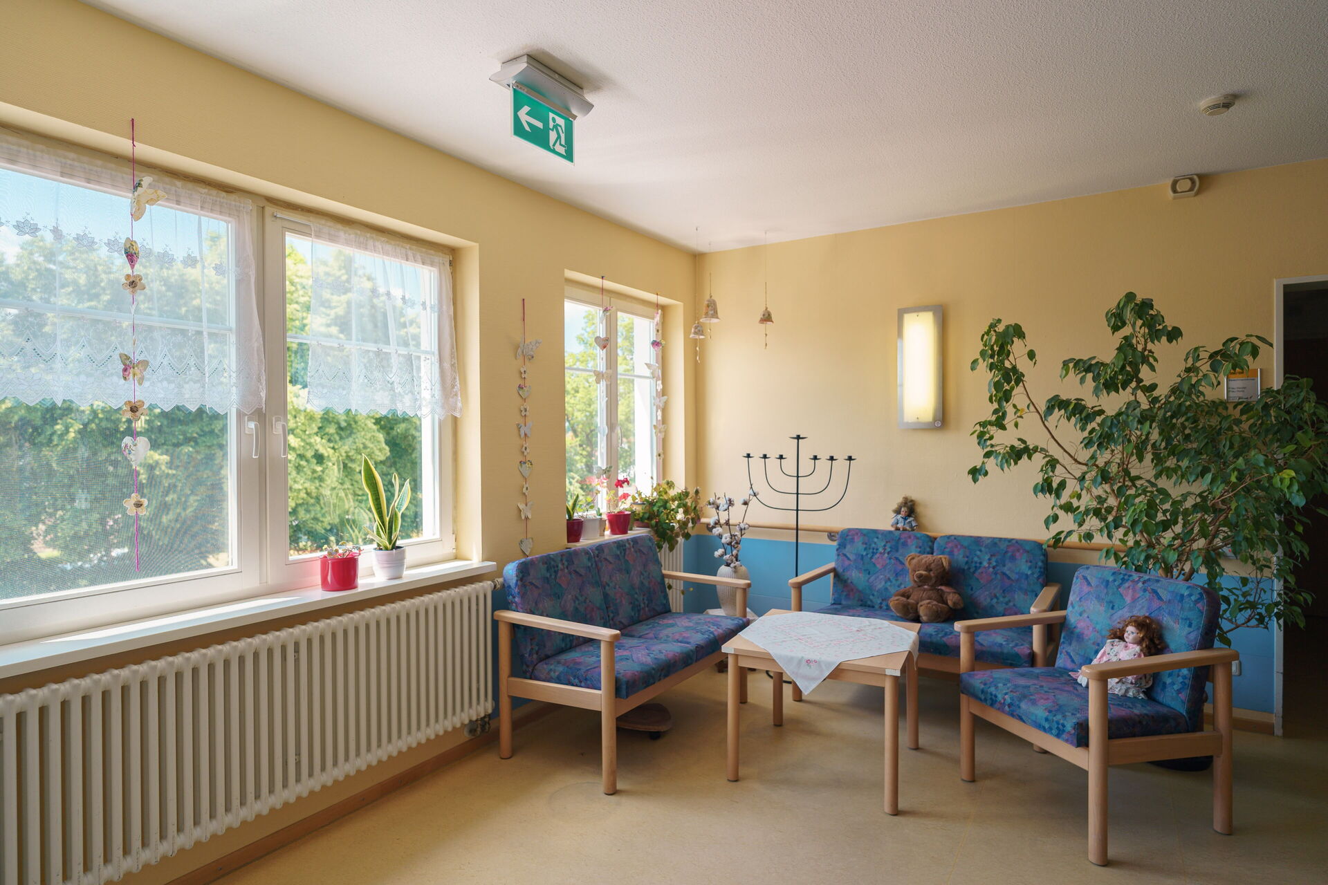 Heller Aufenthaltsraum. Drei Sitzmöbel mit blauen Auflagen und einem kleinen Tisch dazwischen.