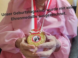 Alte Frau im Rollstuhl trägt eine goldene Medaille. Im Bild schwarzer Text: Geburtstagskind wurde mit Ehrenmedaille ausgezeichnet.