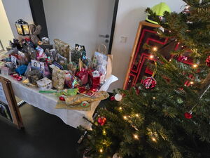 Auf einem Tisch stehen ganz viele bunt verpackte Geschenke. Daneben ist ein mit Lichtern und Kugeln geschmückter Tannenbaum.
