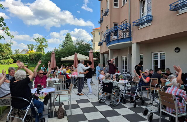 Eine große Terrasse, Stühle, Tische, Menschen in Rollstühlen und mit Rollatoren. Es herrscht ausgelasssene Stimmung, einige schwenken die Arme in der Luft und in der Mitte wird getanzt.