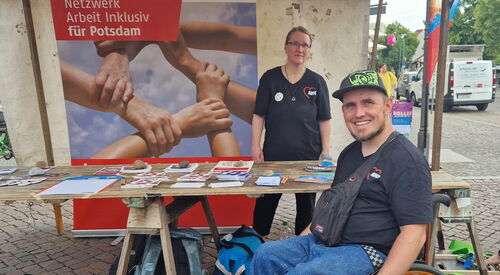 Ein Stand auf dem Fest mit einem Plakat von Netzwerk Arbeit inklusiv für Potsdam. Info-Material auf einem Tisch. Eine Frau im AWO T-Shirt steht hinter dem Tisch. Davor ein Rollstuhlfahrer im Aktivrollstuhl mit AWO T-Shirt und Base Cape.