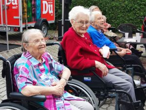 Drei sehr alte Frauen in klobigen Schieberollstühlen nebeneinander geparkt. Die Frau in der Mitte sieht in die Kamera und lächelt leicht. Sie hat weiße Haare, Brille, eine rote Strickjacke und eine graue Stoffhose an.