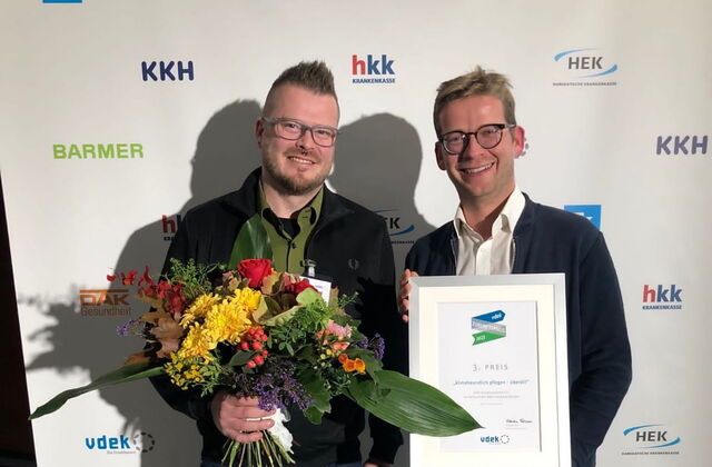 Zwei Männer vor einer Werbewand. Der jüngere Mann hat einem großen Blumenstrauß in der rechten Hand. Der Mann daneben hält eine gerahmte Urkunde zum 3. Platz vor sich.