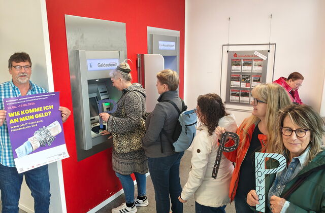 Menschen-Schlange vor Geldautomat. Einige Frauen halten ein großes P In die Kamera. Ein Mann steht links außen und hält ein lila Plakat "Wie komme ich an mein Geld?"