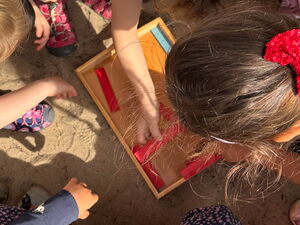 Von oben. Kinder mit einem Holztablett im Sand. Es liegen farbige Holzstäbe im Tablett. Kinder beugen sich darüber.