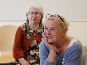 2 Frauen. Frau im Vordergrund lächelt. Hat das Gesicht in die rechte Hand gestüctzt. Sie hat ihre Brille auf den Kopf geschoben und hat kleine Ohrringe. Sie hat ein hellblauen Pulli an.