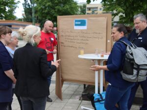 Draußen. 6 Personen stehen um einen Flipchart herum. 4 Politikerinnen unterhalten sich. 2 Männer stehen neben dem Flipshart. Einer im roten T-Shirt hält einen Stift in den Händen.