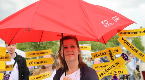Frau mit großem roten AWO-Regenschirm. Im Hintergrund Menschen mit gelben Schildern. Text auf verschiedenen Schilder: Vorleser, Schleifenbinder, Erzähler Dienstplaner, Küchenfee