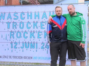 Draußen. Weißes Plakat: „Waschhaus trocken rocken 12. Juni 24“ Die einzelnen Buchstaben und Zahlen sind blau und mit Wäscheklammern an Leinen aufgehängt. Vor dem Plakat stehen zwei Männer in Trainingsanzügen. Eine mit grüer Adidas Jacke und kurzer Hose,.