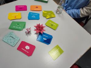 Eine Tischplatte, darauf ein Kartenspiel, die Karten haben verschiedene Farben und sind danach in Stapeln sortiert. 2 Fröbelsterne unterschiedliche Größen liegen in der Mitte.