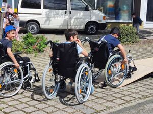 Drei Jungs die in großen unhandlichen Krankenhaus-Rollstühlen sitzen. Eine Junge versucht über eine kleine Rampe zu fahren. Die beiden Anderen folgen ihm.