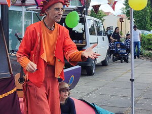 Ein farbenfroh gekleideter Mann mit Narrenmütze bespaßt Kinder und Erwachsene. Er trägt Orange-Rote Kleidung.