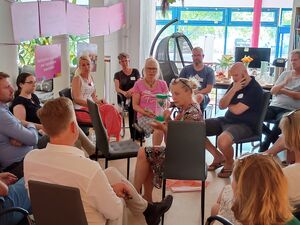 15 Menschen sitzen in zwei Stuhlkreisen. Frau mit mittleren Kreis hält eine grüne Sanduhr durch die grüner Sand rinnt.