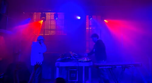 DJs am Pult, es ist dunkel, rot blaues Scheinwerferlicht sorgt für Party-Feeling. Tanzende Menschen im Vordergrund.