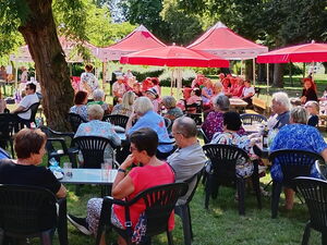 Große Wiese mit Schattenplätzen unter Bäumen und roten Sonnenschirmen. Viele Menschen sitzen draußen.