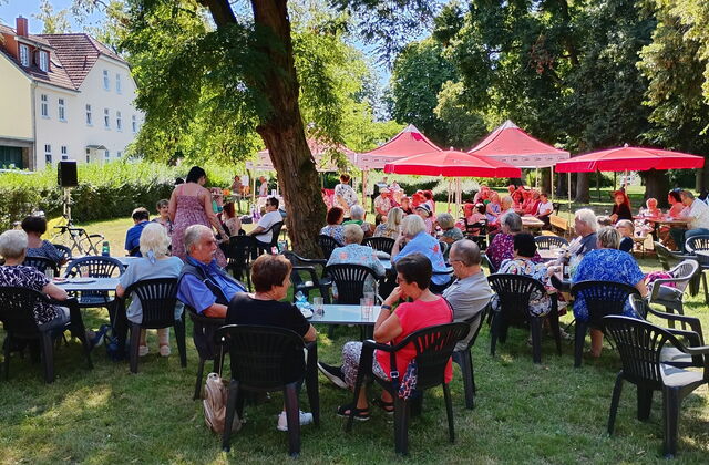 Große Wiese mit Schattenplätzen unter Bäumen und roten Sonnenschirmen. Viele Menschen sitzen draußen.