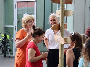 Ein Bild in die Menge. Frauen stehen beieinander und unterhalten sich. Sie sind sommerlich gekleidet. Ein Wegweiser mit Pfeilen in verschiedene Richtungen.