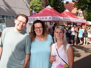 Draußen, Sonne. Ein Mann und zwei Frauen. Mann mit Brille und hellem T-Shirt, Frau lange gewellte Haare, Brille, hellblaues T-Shirt, zweite Frau lange blonde glatte Haare mit Seitenscheitel, weißes ärmelloses Shirt. Lächeln in die Kamera.