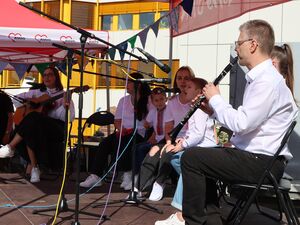 Kleine Bühne, Frau mit Gitarre sitzt auf einem Stuhl. Kinder singen. Mikrofone auf der Bühne und ein Mann in weißem langärmlichen Hemd spielt Klarinette.