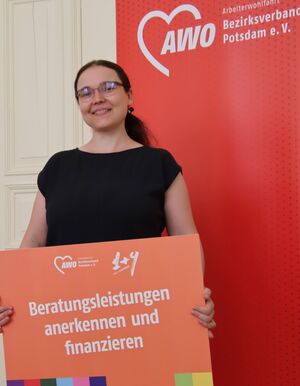 Portrait: Junge Frau mit langen schwarzen Haaren zum Pferdeschwanz gebunden, lächelt fröhlich. Sie träägt ein schwarzes T-Shirt. Sie hält ein großes Schild vor sich. Eine der 1+9 Forderungen. Darauf steht: Berratungsleistungen anerkennen und finanzieren.