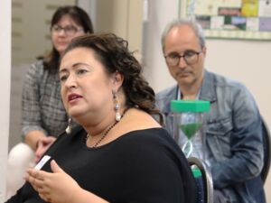 Frau mit langen dunklen Haaren sitzt auf einem Stuhl und spricht. Hinter ihr sitzt ein Mann mit grauen Haaren, Brille, Jeansjacke. Er hat eine grüne Sanduhr auf dem Schoß.