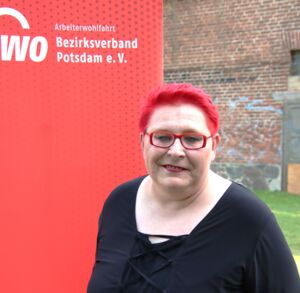 Portrait. Frau lächelt. Sie hat rote kurze Haare, eine rotgerahmte Brille und trägt roten Lippenstift. Sie hat ein schwarzes Oberteil an. Sie stehr draußen vor einem roten AWO Roll-Up-Banner.